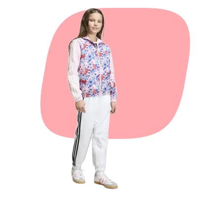Mädchen steht vor rosa Fläche und trägt weiße Trainingshose mit Streifen und bunte Kapuzenjacke