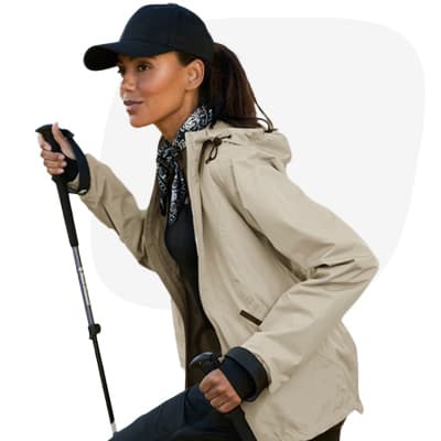 Frau in Outdoorjacke und Basecap wandert mit Stöcken vor rosa Hintergrund