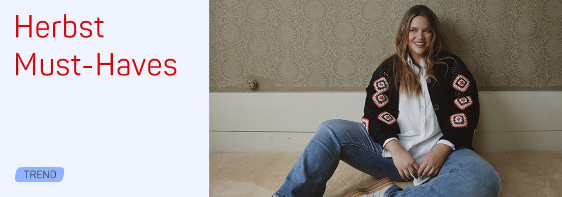 Herbst Must-Haves – sitzende Frau in Jeans und gemusterter Strickjacke lehnt entspannt an der Wand