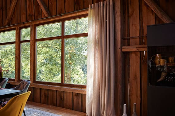 Vorhang vor großer Fensterfront in Holzraum, Blick auf grüne Bäume, daneben Essbereich mit Stühlen und Geschirrvitrine