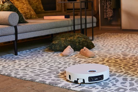 Flacher Saugroboter fährt über gemusterten Teppich im Wohnzimmer vor Sofa, Kissen und Hausschuhen