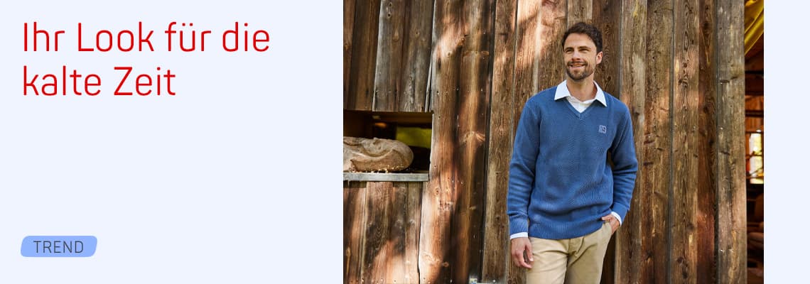Ihr Look für die kalte Zeit – Mann im blauen Pullover lehnt entspannt an Holzwand draußen