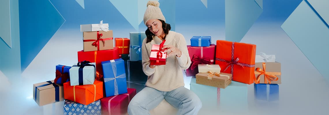 Frau in Mütze und Pullover hält rotes Geschenk, umgeben von bunten, verpackten Geschenken vor blauem Hintergrund.