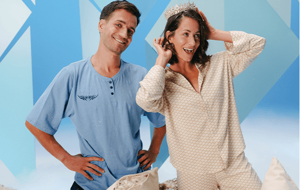 Frau in gemusterter Schlafanzughose und Oberteil trägt Krone, steht neben Mann in blauem T-Shirt vor blauem Hintergrund.