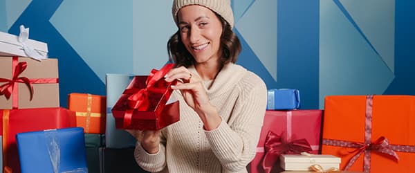Frau in weißem Pullover hält geöffneten Geschenkkarton und Parfümflasche, umgeben von bunt verpackten Geschenken