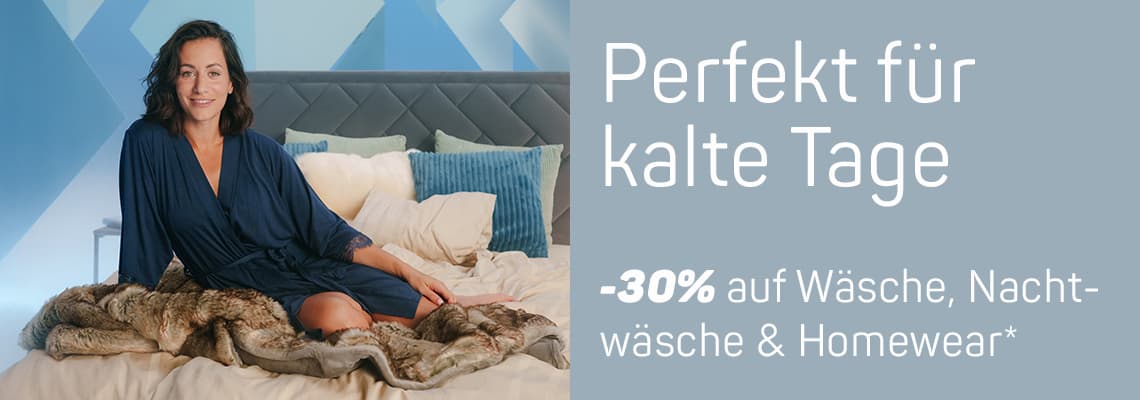 Perfekt für kalte Tage - 30% auf Wäsche, Nachtwäsche & Homewear* – Frau in dunkelblauem Morgenmantel sitzt auf Bett mit Decken