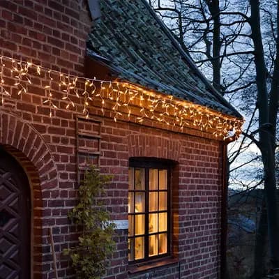 Backstein-Haus mit Lichterkette über Fenster und Tür, beleuchtet bei Dämmerung, Bäume im Hintergrund