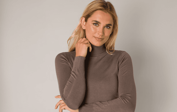 Frau in braunem Rollkragenpullover steht vor neutralem Hintergrund und berührt ihr Gesicht.