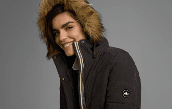 Eine Person in einem dunklen Mantel mit einer pelzbesetzten Kapuze vor einem grauen Hintergrund.