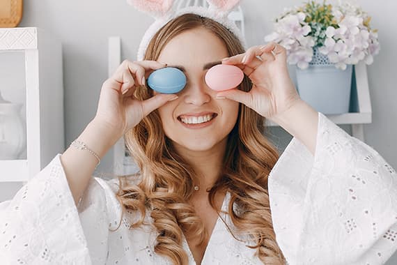 Frau mit langen, lockigen Haaren trägt Hasenohren und hält etwas vor ihr Gesicht. Hintergrund mit Blumen und Regal.